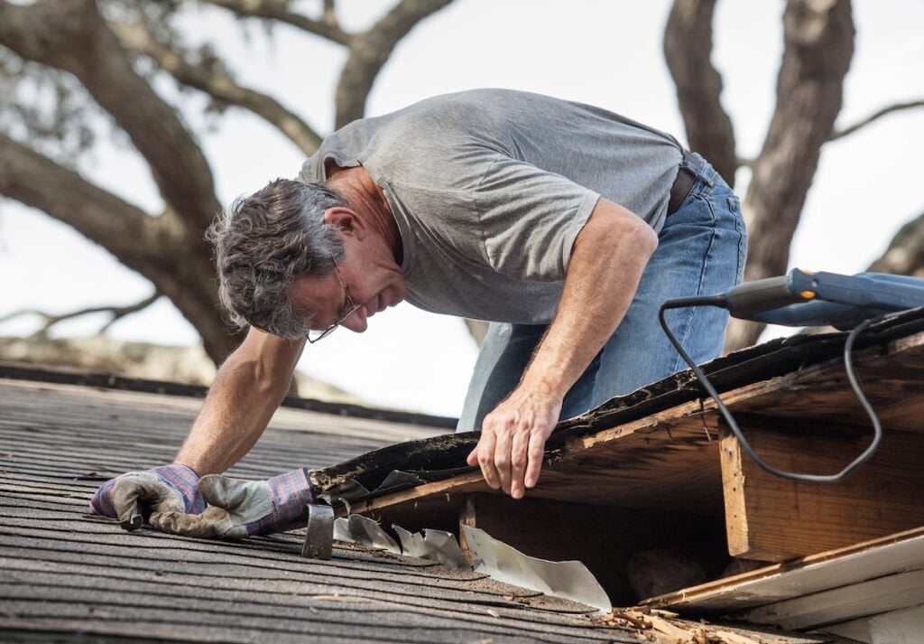 Close up view of man using crowbar and saw to remove rotten wood from leaky roof decking.